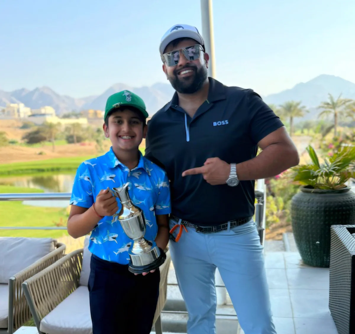 A golfer in a navy polo standing beside a junior golfer in a blue shirt holding a trophy